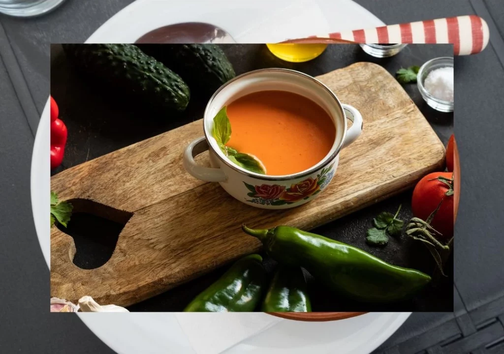 Taza de gazpacho andaluz, sobre una tabla de madera, junto a pimientos y tomates frescos. Pertenece a la imagen de portada del artículo de "Gastronomía: La Receta del Gazpacho de la Subbética.", del blog de casaruralsanantonio.es